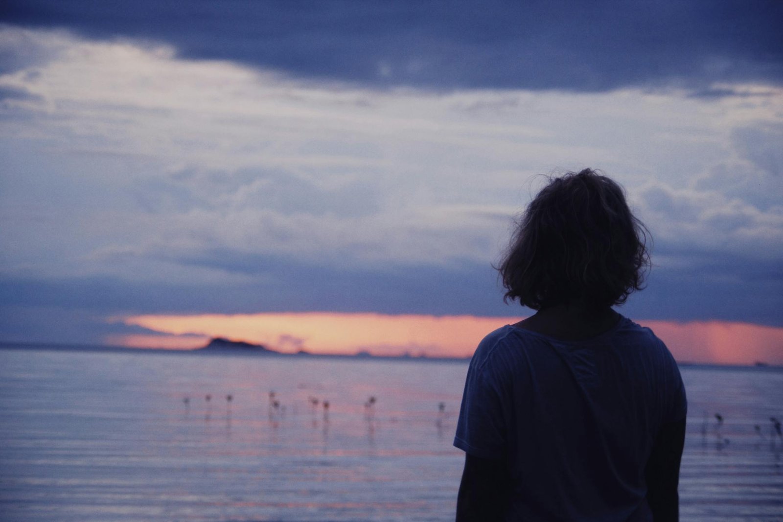 A person stands at the shore, gazing at a vibrant sunset over the calm sea.