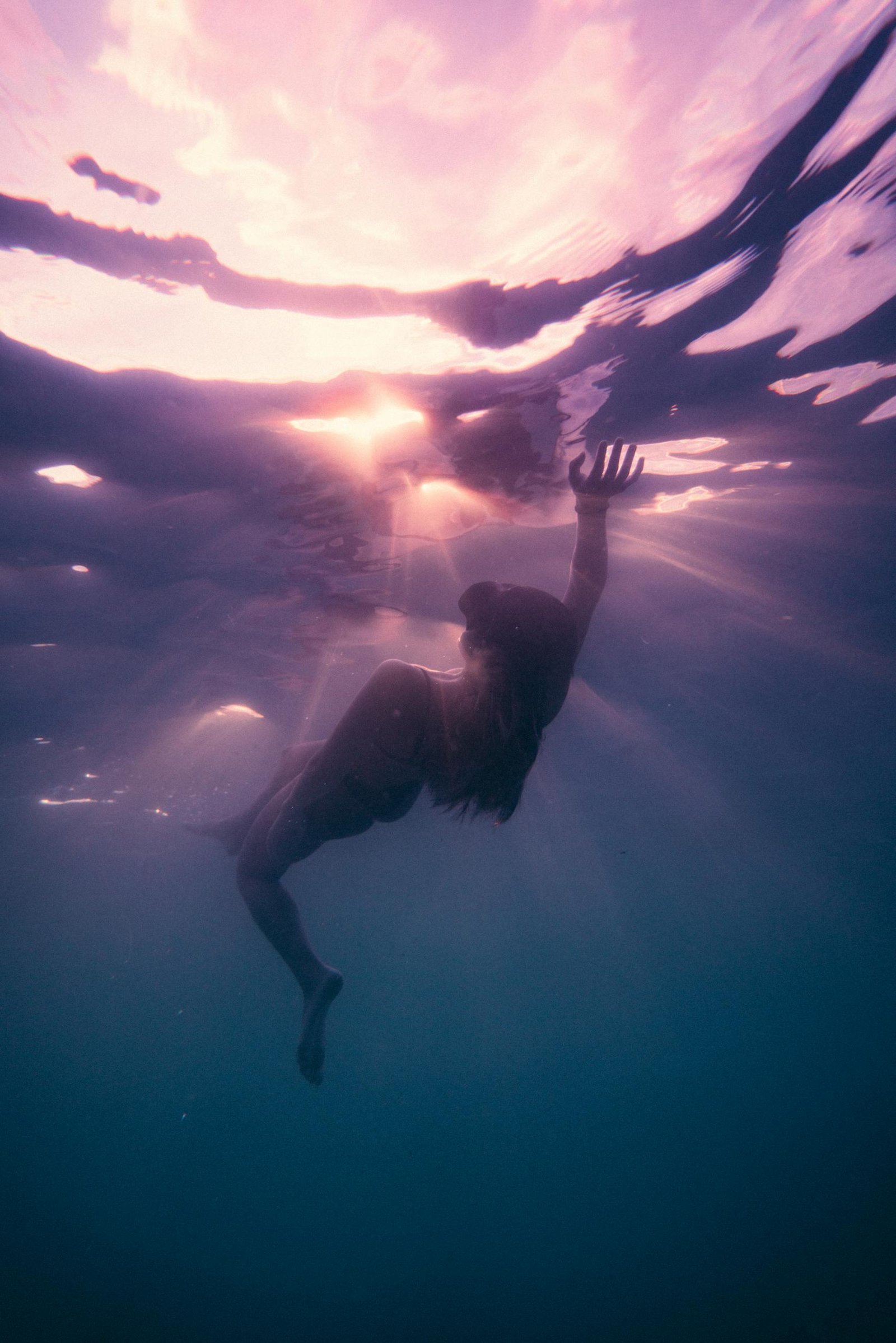 A serene underwater scene with a silhouette of a woman swimming towards the surface during sunset.