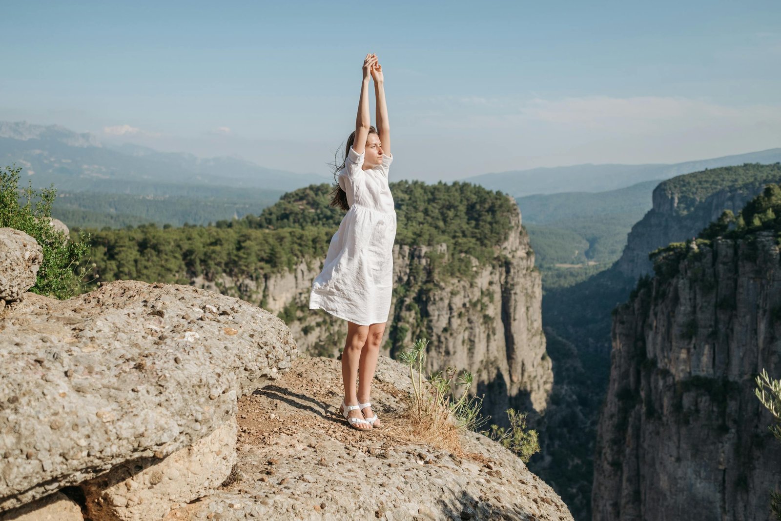 A woman in a white dress stands on a rocky cliff, arms raised, embracing nature's beauty.