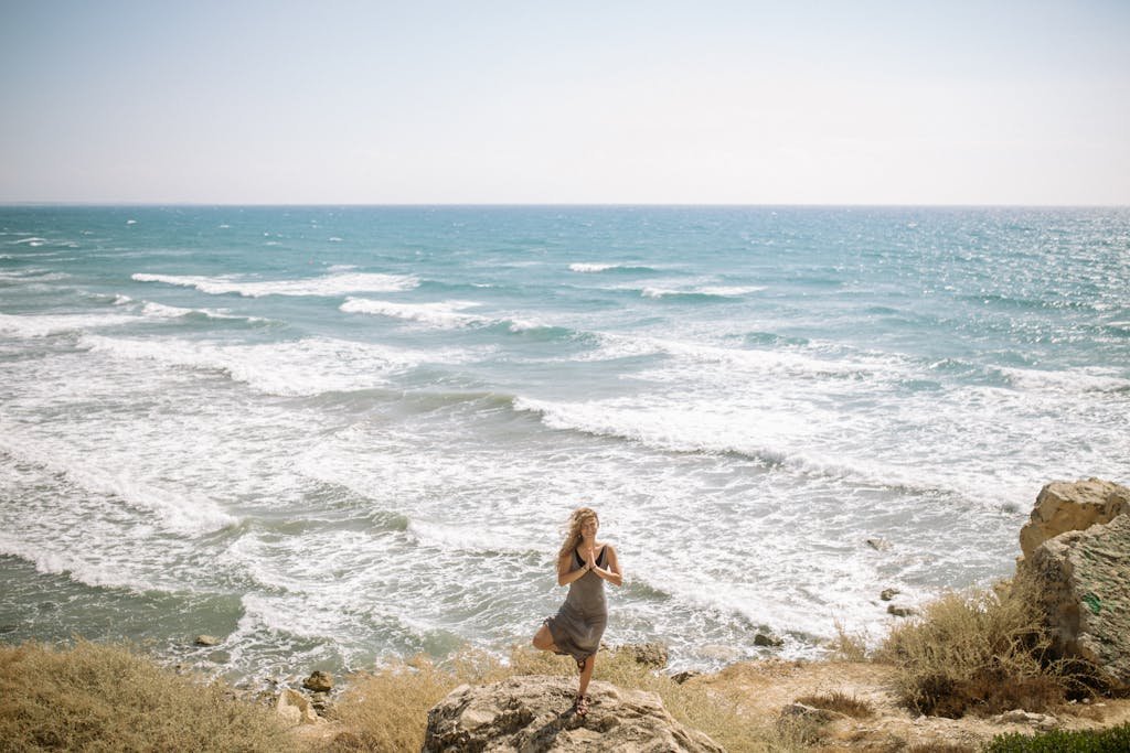 A woman performs a yoga pose on a rocky cliff by the ocean, embracing nature's tranquility.