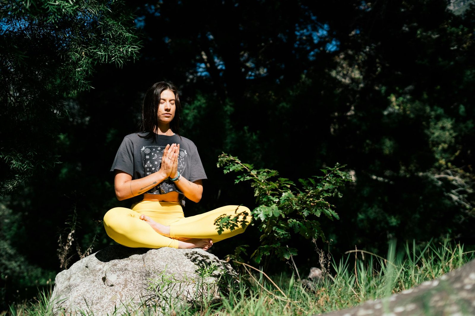 A woman practicing yoga outdoors, seated in a meditation pose on a rock, surrounded by lush greenery.