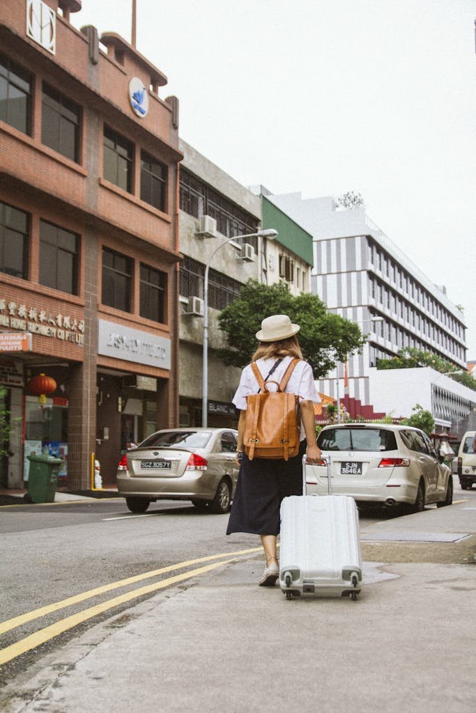 A woman with a suitcase and backpack walks down a busy city street on a sunny day.