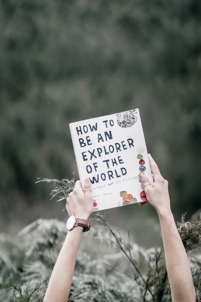 Book titled 'How to Be an Explorer of the World' held by hands against nature background.