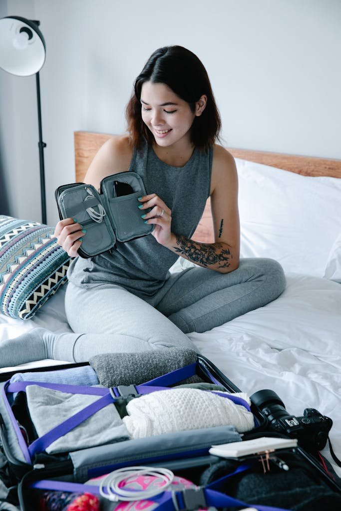 Woman in gray outfit packing suitcase on bed in well-lit room, smiling at electronic organizer.