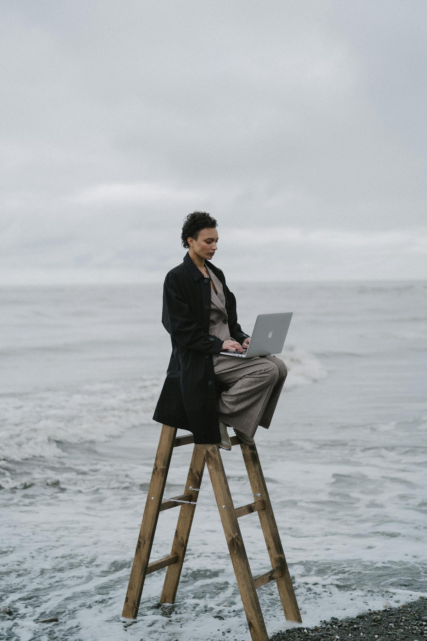 Woman sitting on a wooden ladder by the ocean, typing on a laptop, embodies remote work outdoors.