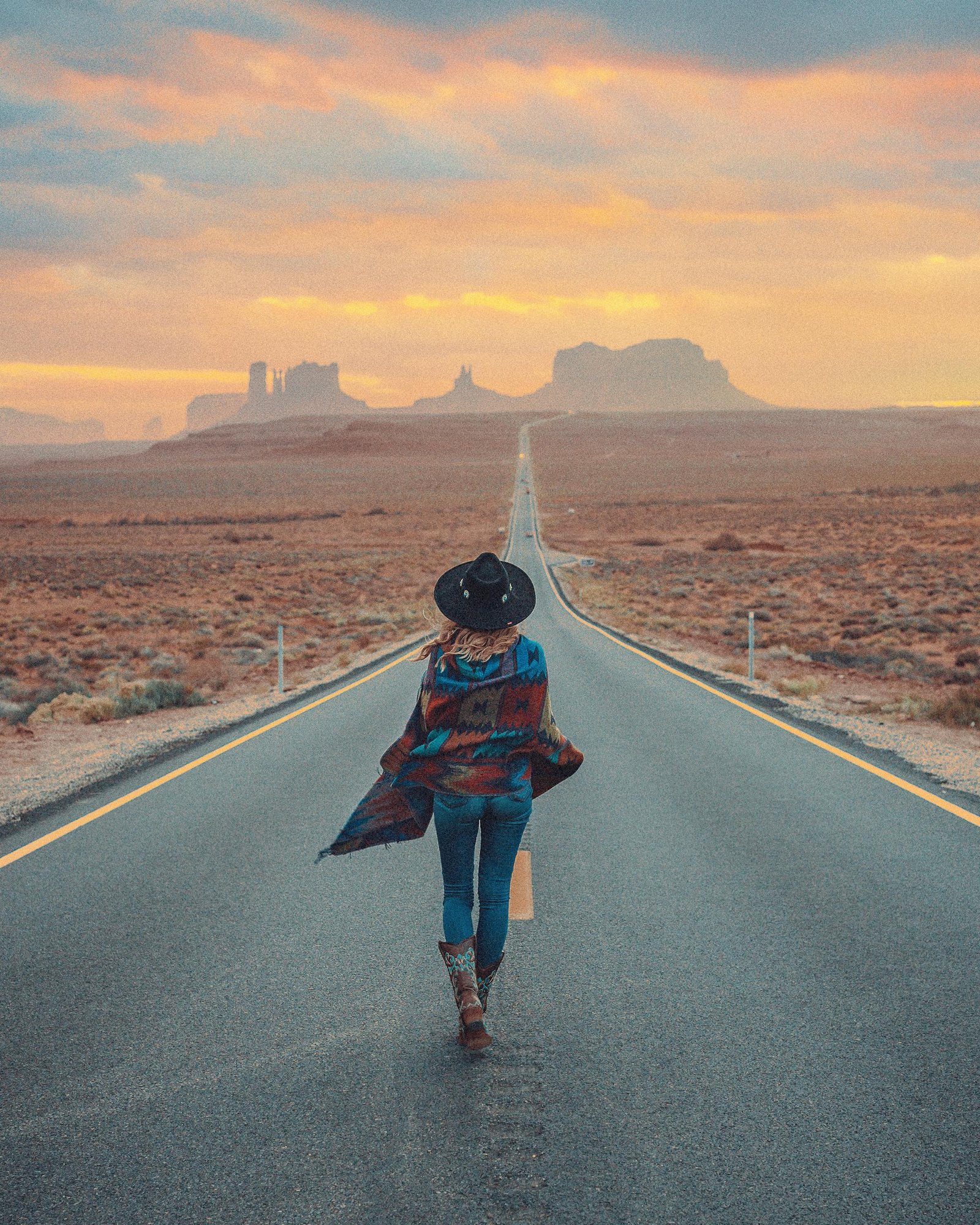 Woman walking on a desert road towards Monument Valley under a dramatic sunset sky.