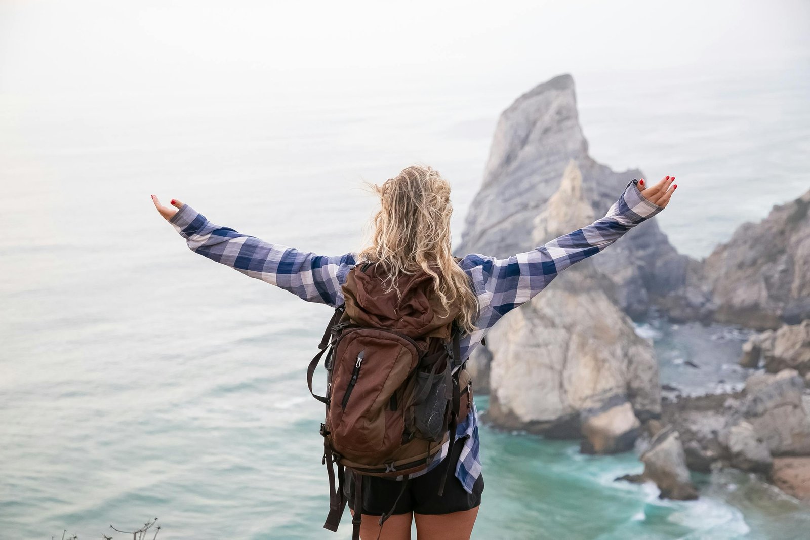 Woman with backpack stands on mountainside with arms wide open, overlooking scenic ocean view.