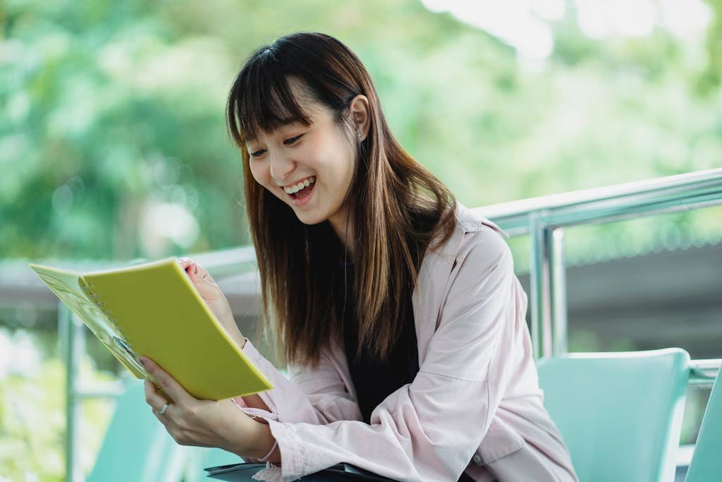 Young happy ethnic female student watching exercise book while sitting on chair in daylight