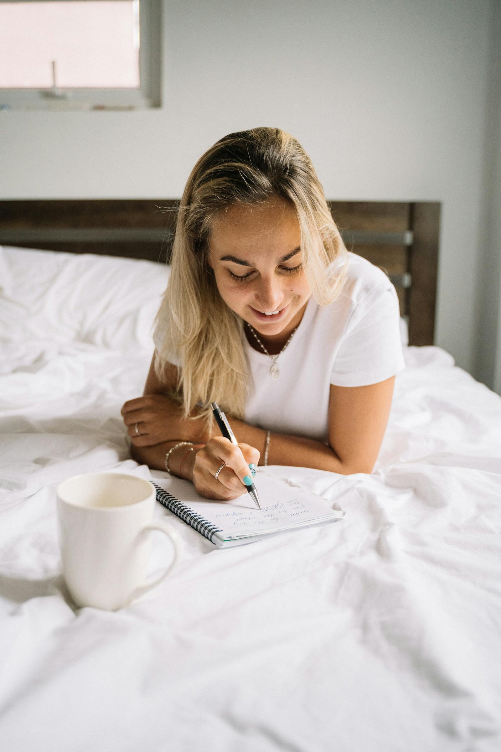 Young woman comfortably writing in a notebook on a cozy bed with a mug nearby.